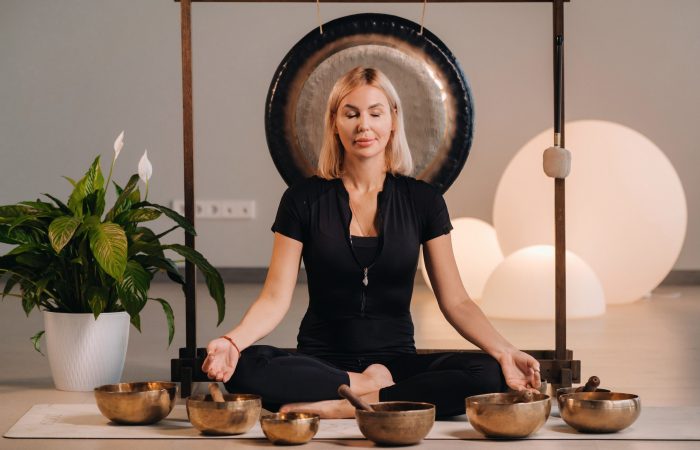 A woman sits in a lotus position next to Tibetan bowls, sitting on a yoga mat against the background of a gong.